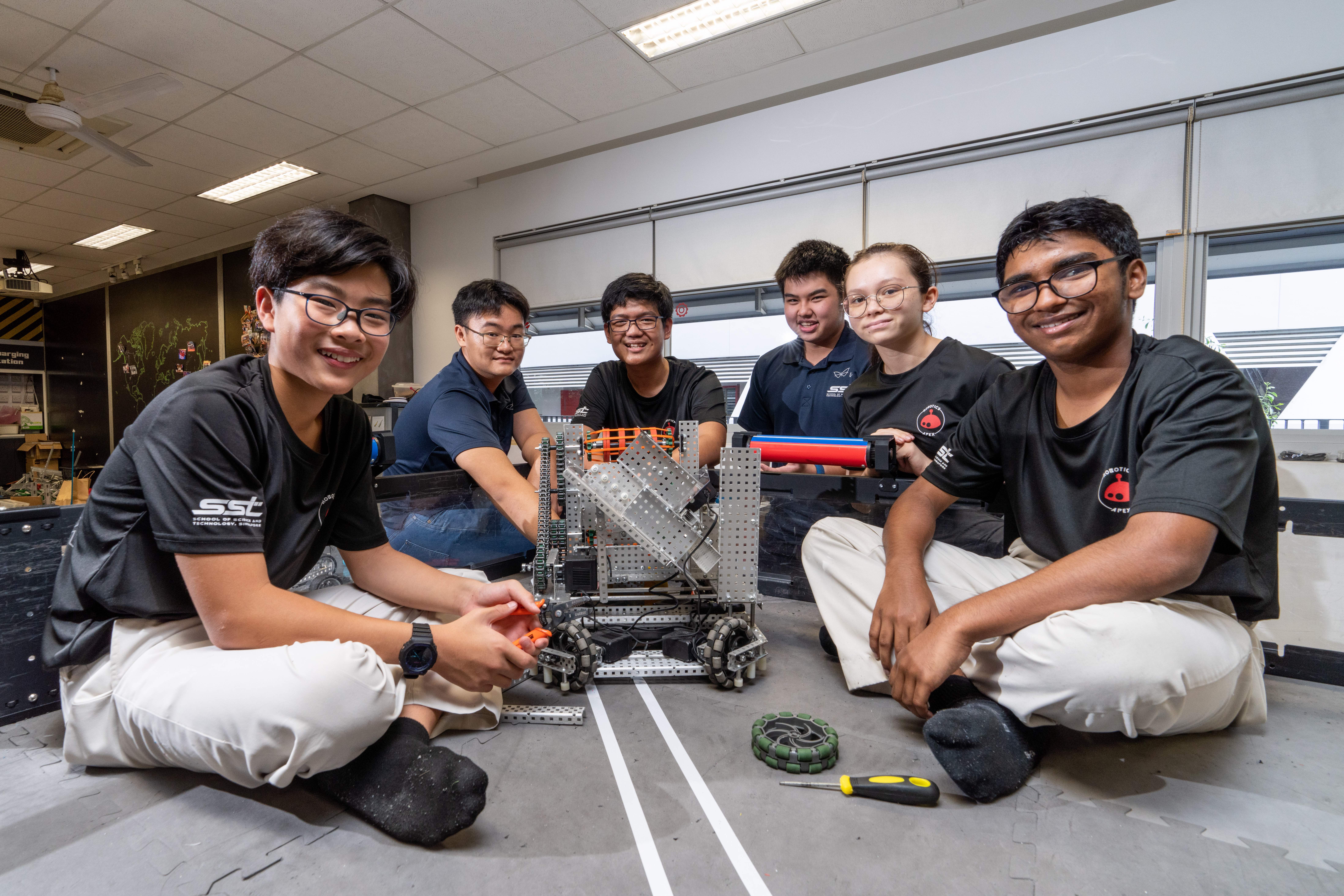 Six students sit on a lab floor around a metal robot, smiling, with tools and wheels nearby.